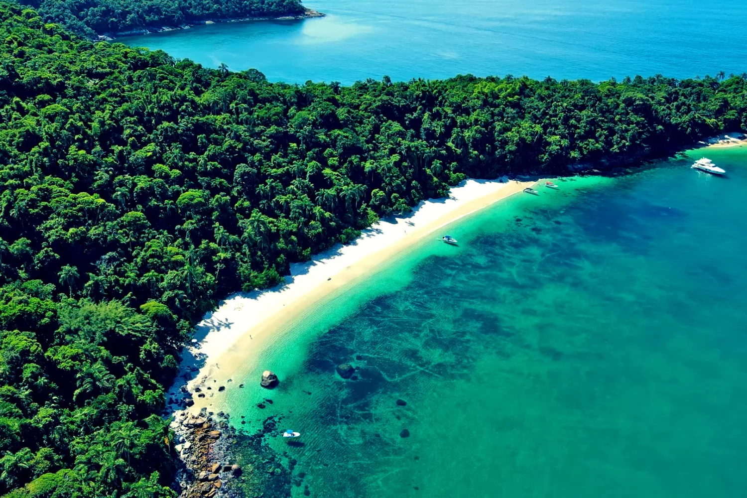 Visão aérea em perspectiva, mais aproximada, da Praia de Jurubaíba em Angra dos Reis/RJ, mais conhecida como Praia do Dentista. O verde da vegetação exuberante abraça a faixa de areias brancas, combinando com o verde cristalino do mar.