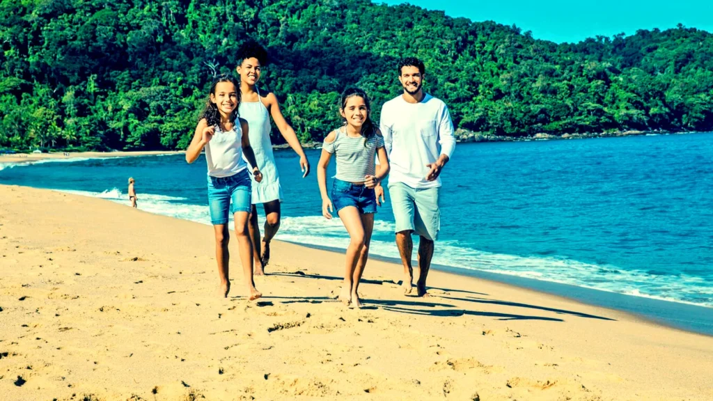 Quatro pessoas da mesma família: pai, mãe, duas filhas entre 9 e 10 anos, caminhando na praia, na faixa de areia próximo ao mar, em dia de sol com a marola espumando na parte direita.